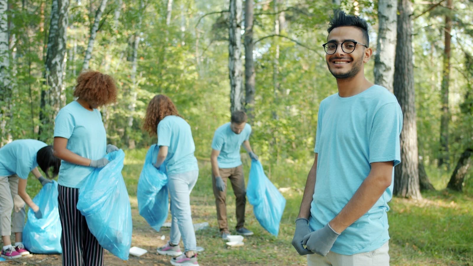 A smiling young man in work gloves stands in a sunny wood while other young volunteers in matching T-shirts fill bags with litter behind him.