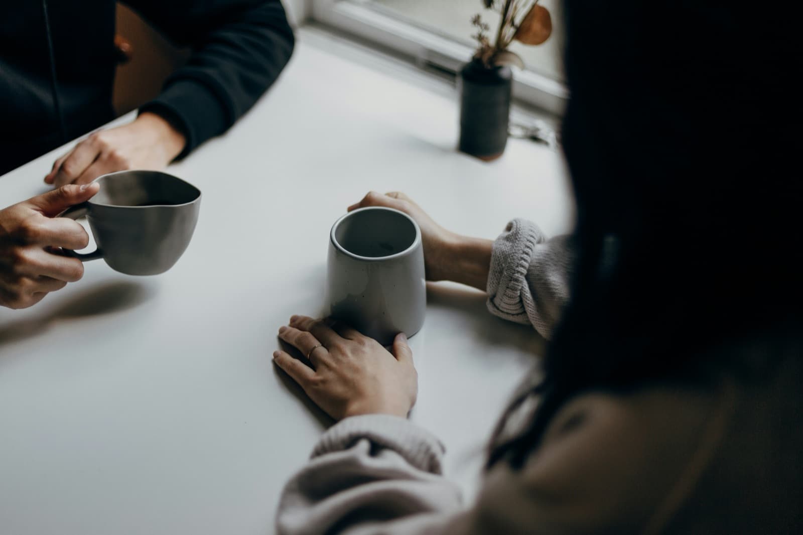 Two people's hands around coffee mugs on a white table by a window, mid-conversation.